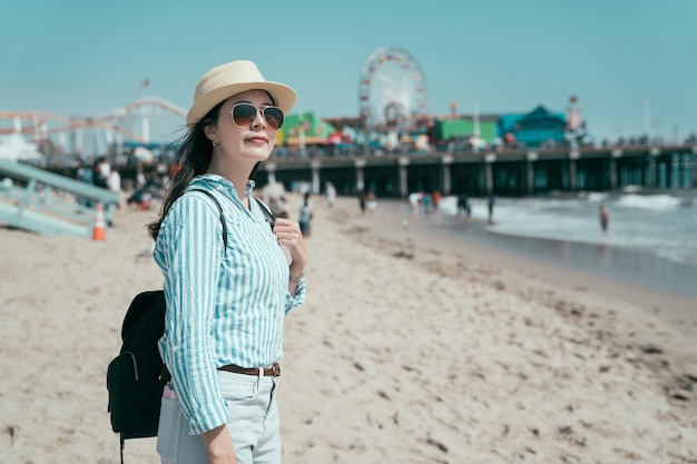 lado feliz sonriente joven viajero en sombrero de paja y gafas de sol con mochila. elegante turista de pie en la playa de arena disfruta de una hermosa vista al mar. bokeh del parque de atracciones bajo el cielo azul.