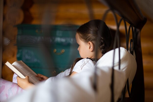 Lado joven de la muchacha capturada detrás de la cama acostado en la almohada sosteniendo el libro en las manos mirando a la bo...