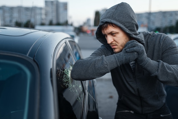 Ladrão de carros quebrando vidros de portas, trabalho criminoso, ladrão. ladrão masculino com capuz abrindo veículo no estacionamento. roubo de automóveis, crime automobilístico, vandalismo