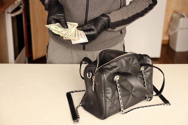 Ladrão de roupa preta e luvas vê em bolsa de mulher roubada aberta O ladrão tira notas de dólar americano de uma bolsa de mulher na cozinha