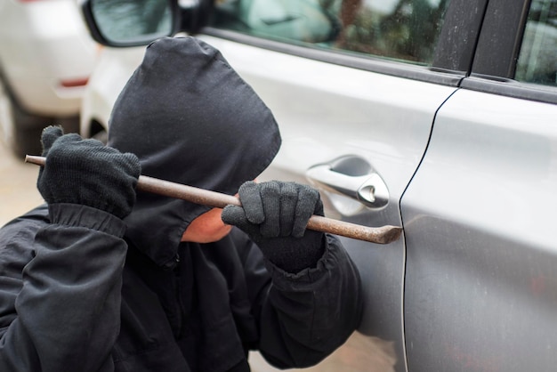 Ladrón con chaqueta con capucha negra usando una palanca para romper la cerradura y robar el vehículo