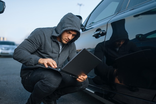 Ladrón de coches con sistema de alarma de piratería portátil, estilo de vida criminal. Vehículo de apertura de ladrón macho encapuchado en el estacionamiento. Robo de automóviles, delitos de automóviles