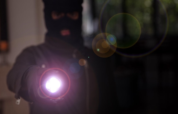 Foto un ladrón enmascarado entrando en una casa con una linterna en un fondo oscuro.