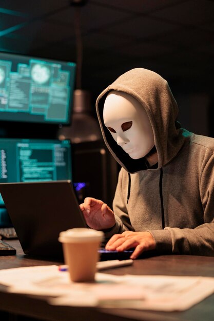 Ladrón enmascarado peligroso instalando virus para hackear el sistema en la computadora, trabajando tarde en la noche en la oficina. Ciberterrorista y criminal con servidor de red de piratería de máscaras y estafa o fraude.
