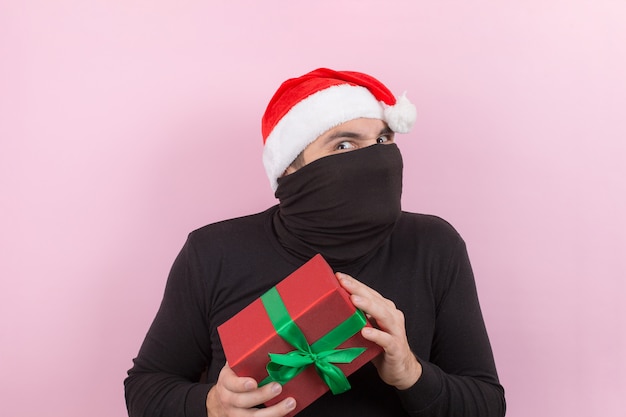 Un ladrón con un sombrero rojo robó los regalos de Navidad de otra persona. Carácter enojado, emociones humanas negativas. Fondo rosa, espacio de copia.