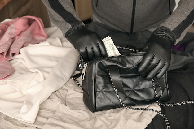 Ladrón con traje negro y guantes ve en una bolsa de mujer robada abierta El ladrón saca billetes de dólares estadounidenses del bolso de una mujer en la cocina