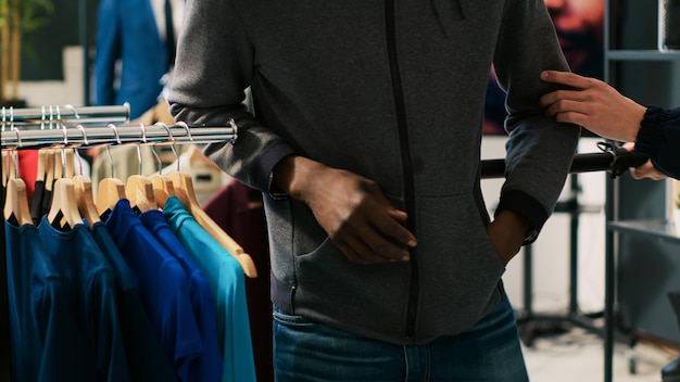 Ladrón tratando de robar una camisa de moda en una boutique moderna, con capucha y gafas de sol para no ser reconocido. Guardaespaldas asiático amenazando a un ladrón afroamericano con un bastón de seguridad en una tienda de ropa