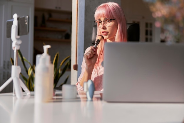 Lady blogger, novo tutorial de tiro com pincel de maquiagem na mesa com produtos cosméticos e laptop