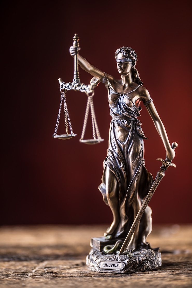 Foto lady justicia sosteniendo espada y figura de bronce a escala sobre mesa de madera.