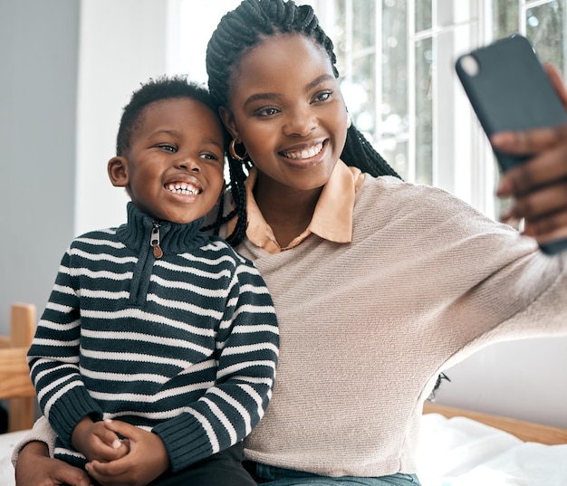 Lächeln Schnappschuss einer attraktiven jungen Frau und ihres entzückenden Sohnes, die Selfies machen, während sie zu Hause auf einem Bett sitzen