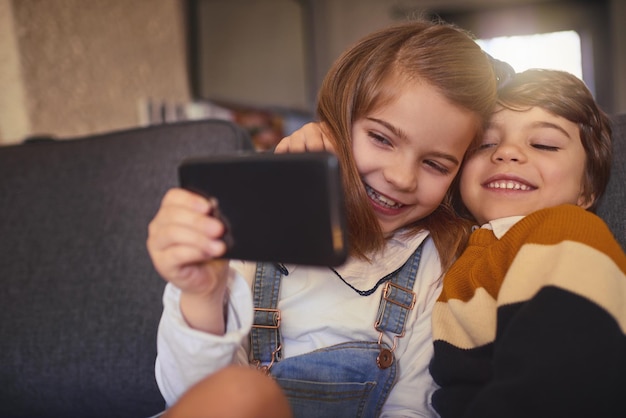 Lächeln Schnappschuss eines entzückenden kleinen Mädchens und ihres jüngeren Bruders, die Selfies machen, während sie zu Hause auf dem Sofa sitzen