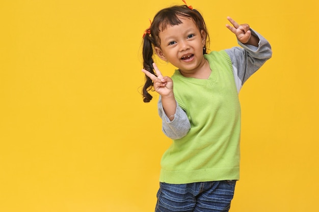 Lächeln Sie süß von einem asiatischen kleinen Mädchen, das Vsign auf gelbem Hintergrund mit Kopierraum zeigt