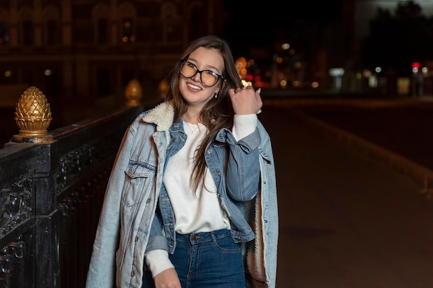 Lächelnd attraktive Brünette Frau in Denim-Kleidung und Brille am Abend Stadt Hintergrund Stilvolles Mädchen in der Nachtstadt