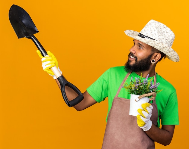 Lächelnd aussehender junger Gärtner afroamerikanischer Mann mit Gartenhut und Handschuhen, der Spaten mit Blume im Blumentopf hält