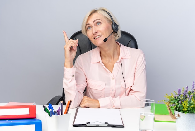 Lächelnd nachschlagende junge Callcenter-Betreiberin mit Headset am Tisch sitzend mit Bürowerkzeugen zeigt nach oben isoliert auf weißer Wand