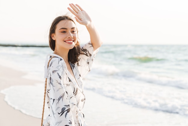 lächelnd optimistisch glückliche junge schöne Frau am Strand zu Fuß an einem schönen sonnigen Morgen zu jemandem zuwinken.