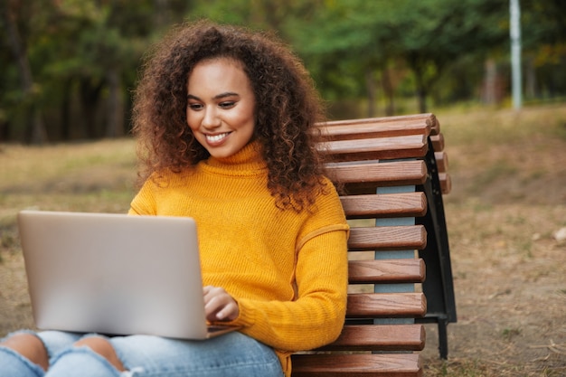 lächelnd schöne junge lockige Frau sitzen im Park im Freien mit Laptop-Computer.