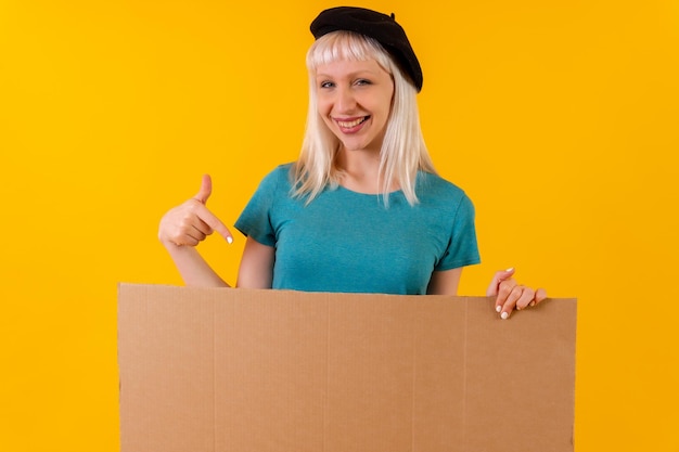 Lächelnd und auf leeres Pappwerbeplakat zeigend blondes kaukasisches Mädchen im Studio auf gelbem Hintergrund