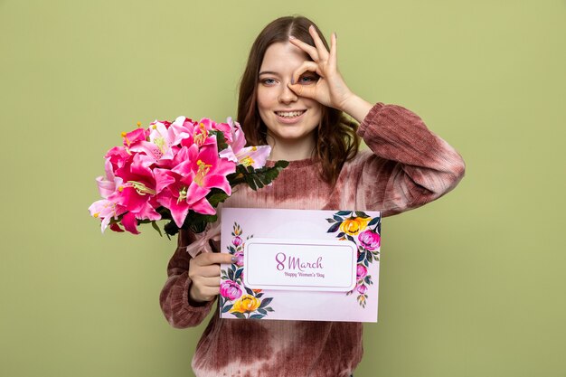 Lächelnd zeigender Blick Geste schönes junges Mädchen am Tag der glücklichen Frau mit Blumenstrauß mit Postkarte isoliert auf olivgrüner Wand