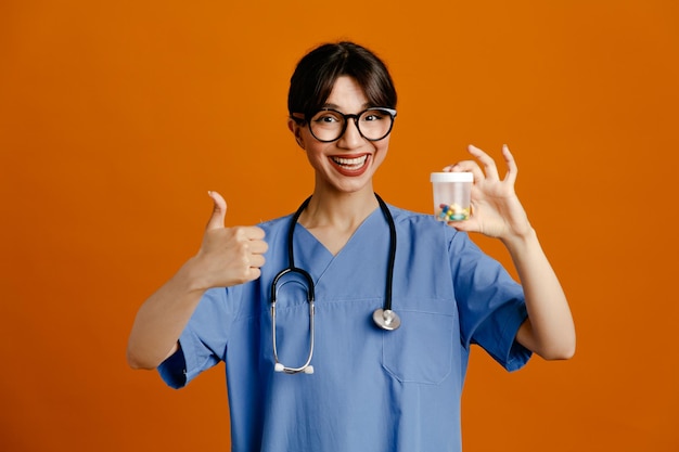 Lächelnd zeigt daumen nach oben hält pillenbehälter junge ärztin mit einheitlichem fith-stethoskop isoliert auf orangefarbenem hintergrund