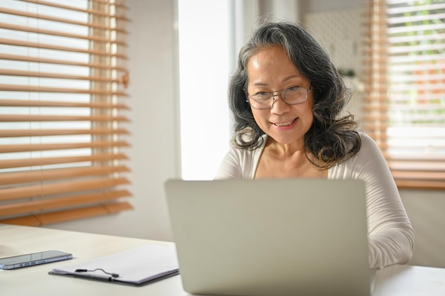 Lächelnde 60-jährige asiatische Geschäftsfrau, die an ihren geschäftlichen Aufgaben auf einem Laptop arbeitet