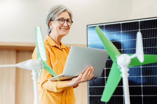 Lächelnde ältere Frau benutzt einen Laptop in der Nähe von Solarpanel und Windmühle Alternative Windenergie