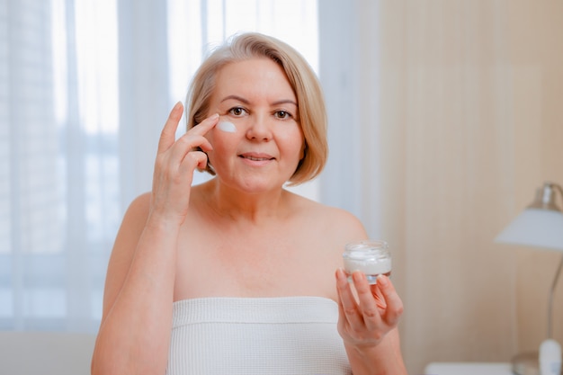 Lächelnde ältere Frau, die Anti-Aging-Lotion anwendet, um dunkle Ringe unter den Augen zu entfernen.