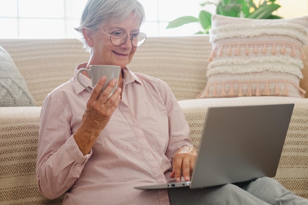 Lächelnde ältere Frau, die zu Hause auf dem Boden sitzt, während sie auf dem Laptop surft und eine Tasse Kaffee hält, ältere Dame, die Technologie und Kommunikation genießt