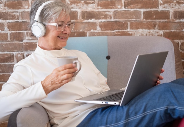 Lächelnde ältere Frau, die zu Hause mit Laptop-Computer auf einem Sessel sitzt und eine Tasse Tee oder Kaffee trinkt. Backsteinmauer Hintergrund