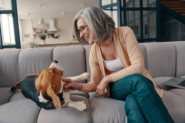 Lächelnde ältere Frau in Freizeitkleidung, die Zeit mit ihrem Hund verbringt, während sie zu Hause auf dem Sofa sitzt