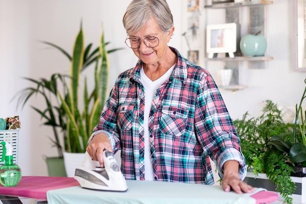 Lächelnde ältere Frau in kariertem Hemd und Brille, die zu Hause Kleidung auf dem Bügelbrett bügelt