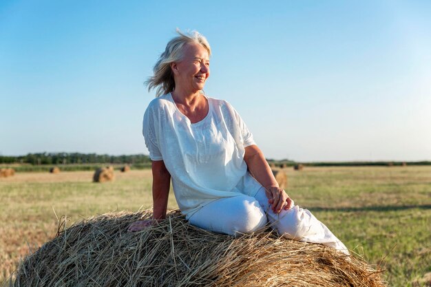 Lächelnde ältere Frau in weißem Pullover und Hose auf einem Feld mit Heurollen bei Sonnenuntergang. Freiheit in der Landwirtschaft, Aktivität in der Natur und Langlebigkeit