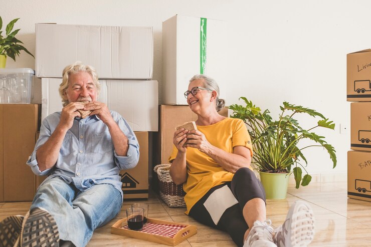 Lächelnde ältere Menschen, weißes Haar, die in der neuen leeren Wohnung mit Umzugskartons arbeiten, auf dem Boden sitzen und Pause machen - Konzept aktiver älterer Menschen und Neuanfang wie im Ruhestand