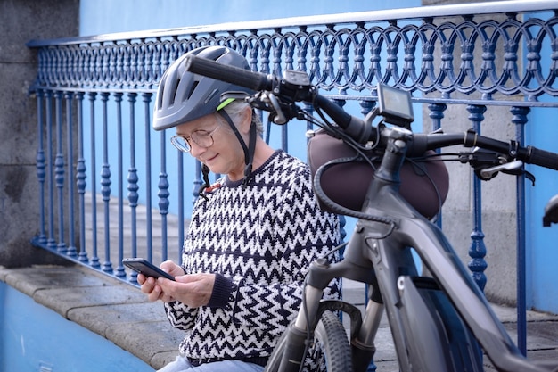 Lächelnde ältere Radfahrerin mit Fahrradhelm, die in der Nähe ihres Fahrrads sitzt und ihr Handy benutzt