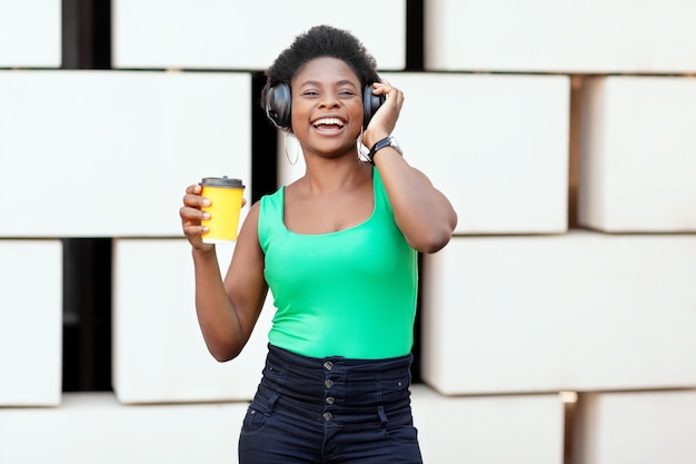 Lächelnde Afrikanerin hält Kaffee in einem Einwegbecher, hört eine angenehme Melodie in Kopfhörern und tanzt.