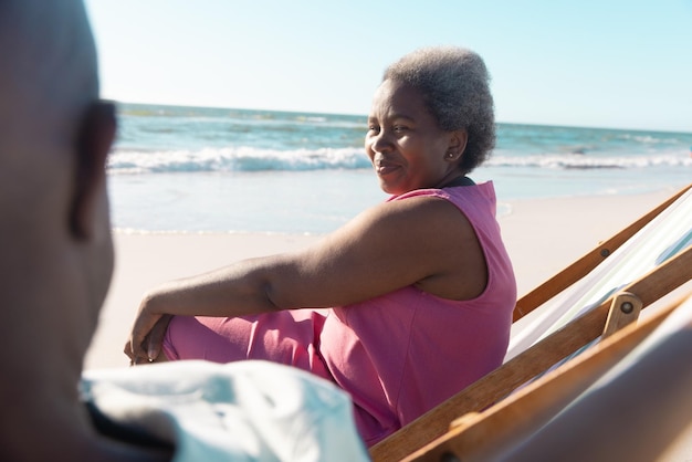 Lächelnde afrikanisch-amerikanische ältere Frau, die mit einem Mann spricht, während sie sich auf Liegestühlen am Strand entspannt.