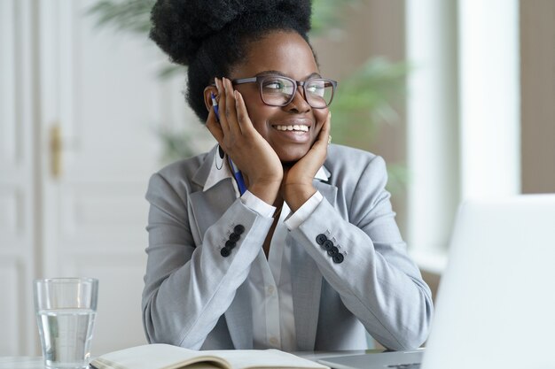 Lächelnde Afro-Frau im Blazer tragen Brille, die am Laptop-Computer zu Hause arbeitet und wegschaut.