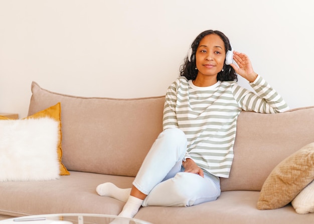 Lächelnde afroamerikanische Frau, die Musik über Kopfhörer hört, während sie auf der Couch sitzt