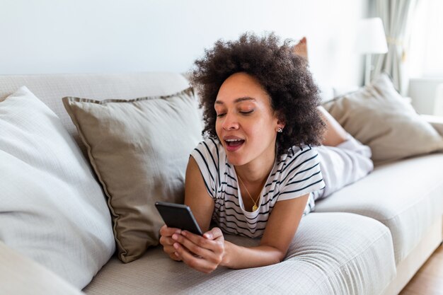 Lächelnde afroamerikanische Frau, die Nachricht auf ihrem Handy liest, während sie auf Sofa zu Hause sitzt.