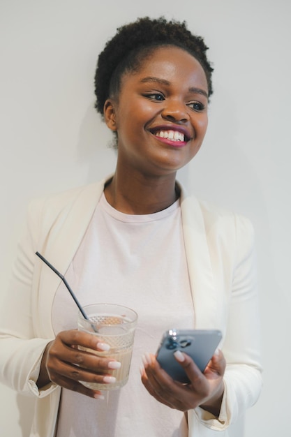 Foto lächelnde afroamerikanische geschäftsfrau in weißer jacke trinkt eiskaffee mit telefon im internetcafé job in der geschäfts- und finanzbranche selbständige frau genießt ein getränk und entspannt sich