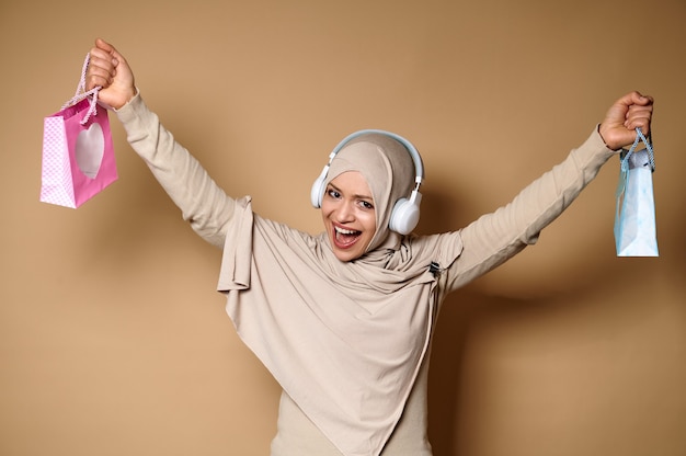 Lächelnde arabische muslimische Frau im Hijab und in den Kopfhörern, die farbige Papiertüten halten und Glück ausdrücken, das in die Kamera schaut
