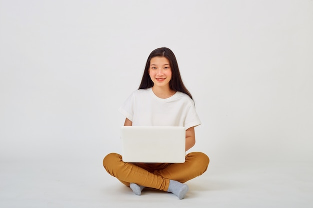 Lächelnde asiatische Frau, die an einem Laptop arbeitet, während sie mit gekreuzten Beinen auf dem Boden sitzt, isoliert auf weißem "Hintergrund, Zurück zum Schulkonzept"