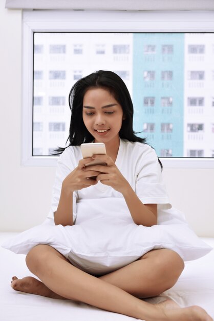 Lächelnde asiatische Frau, die den Handy sitzt auf Bett verwendet