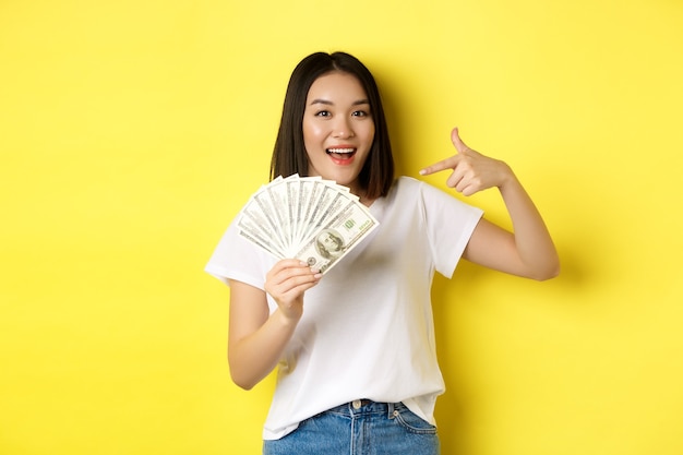 Lächelnde asiatische Frau, die Hände auf Geld zeigt, Dollar zeigt und über gelbem Hintergrund steht.