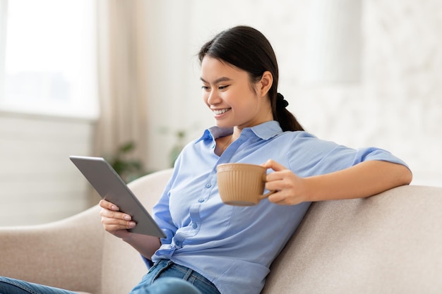 Lächelnde asiatische Frau, die Kaffee trinkt und digitale Tablette verwendet