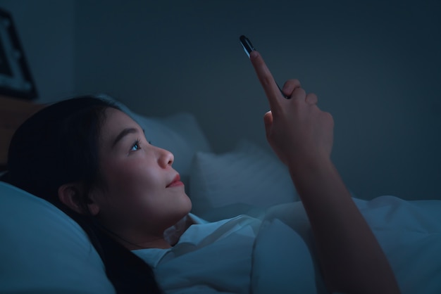 Lächelnde asiatische Frau, die nachts auf dem Bett liegt und Smartphone online sozial verwendet.