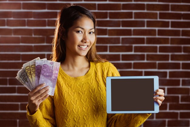 Lächelnde asiatische frau, die tablette und banknoten auf backsteinmauer zeigt