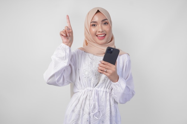 Foto lächelnde asiatische frau mit hijab und weißem kleid benutzt ein smartphone, während sie auf den kopierraum oben zeigt und über einem isolierten weißen hintergrund steht.