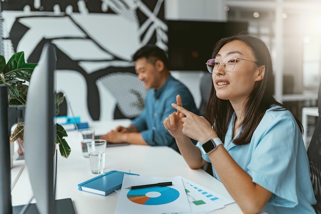 Lächelnde asiatische Geschäftsfrau, die am Computer arbeitet und im Coworking auf dem Hintergrund der Kollegen sitzt