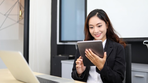 Lächelnde asiatische Geschäftsfrau, die ein Tablet für die Arbeit verwendet, und ein süßes Mädchen, das nach Daten sucht und sich auf ihr Tablet in ihrem Büro konzentriert